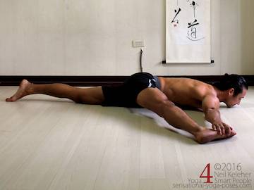 Adductor stretches: prone big toe pose adductor stretch. Lying prone with one leg reaching out to the side with knee pointing forwards. Same side hand is grabbing the blade of the foot. Chin is on the floor with gaze directed forwards and slightly downwards. Neil Keleher. Sensational Yoga Poses.