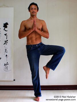 Modified tree pose position balancing on one foot with the other knee lifted to the side but foot is not touching the supporting leg. Hands are in prayer position. Neil Keleher in Blue jeans. Sensaional Yoga Poses.